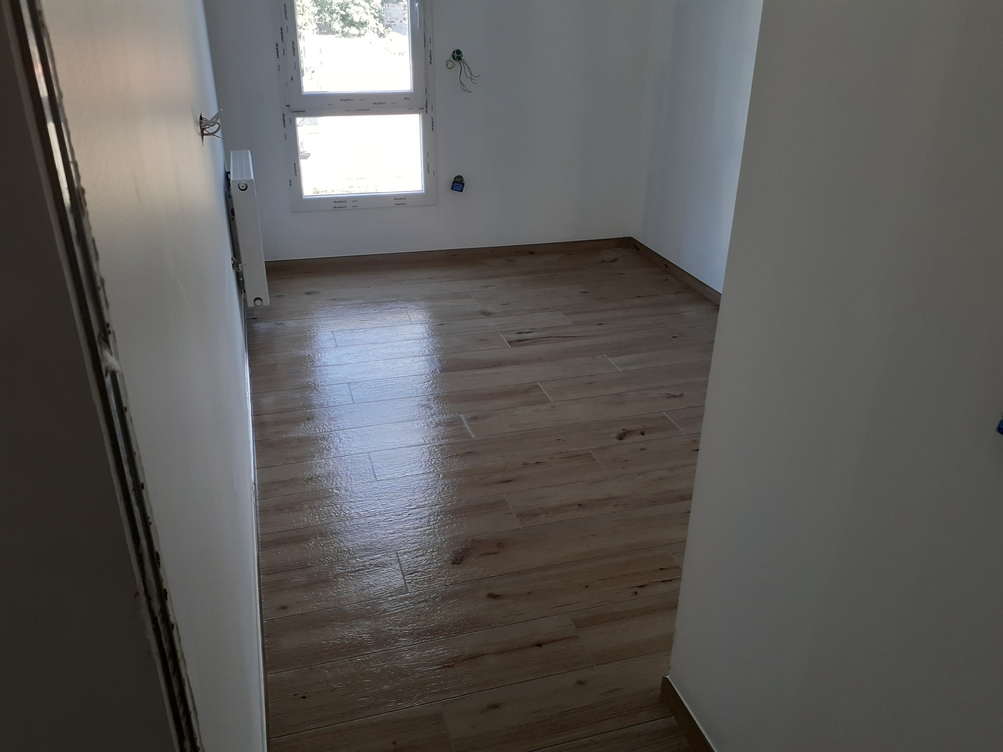 Jasne, nowocześnie wykończone wnętrze z drewnopodobną podłogą z płytek, białymi ścianami, grzejnikiem i widocznymi przewodami elektrycznymi przy oknie.