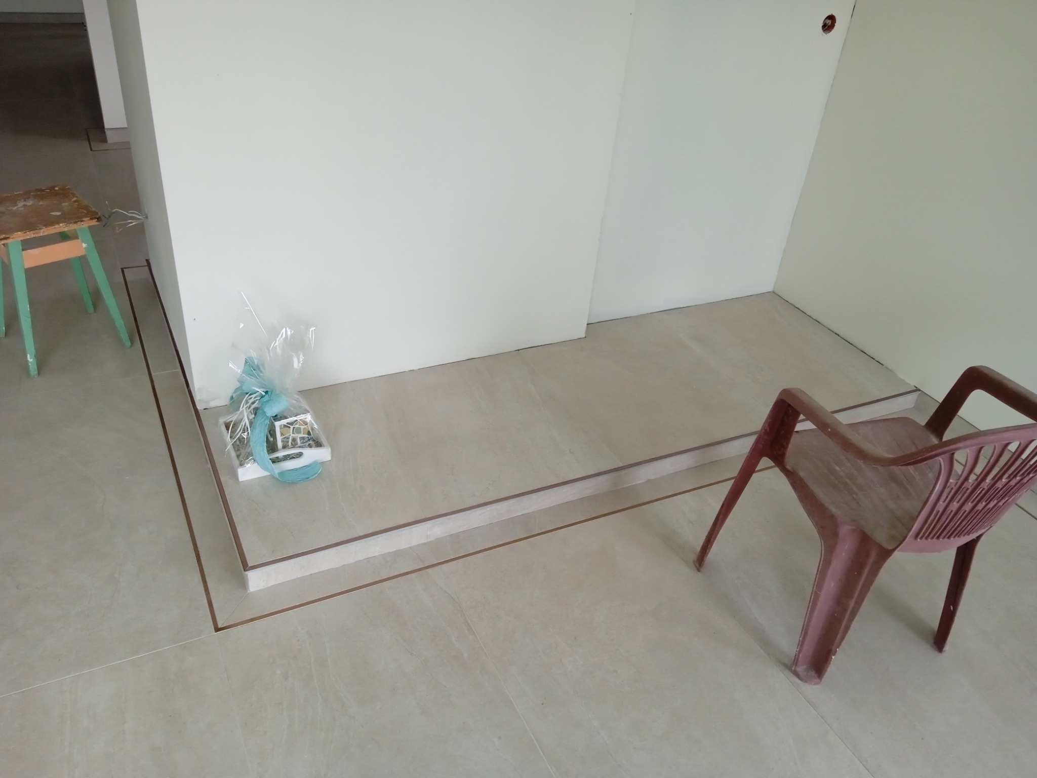 Beżowe płytki podłogowe z dekoracyjnym obramowaniem, prezent w celofanie, plastikowe krzesło i drewniany taboret w surowym wnętrzu podczas prac wykończeniowych.