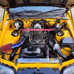 PANFAB Wiktor Drążek - Żółty samochód Toyota Supra z otwartą maską, ukazujący zmodyfikowany silnik z czerwonym filtrem powietrza i niebiesko-fioletowym dolotem, oraz rozpórką kielichów w kolorze czerwonym.