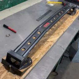 Czarny, metalowy element konstrukcyjny z otworami i logo, leżący na drewnianej desce na metalowym stole warsztatowym, obok narzędzi i markera.