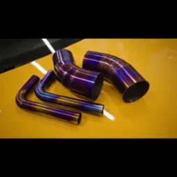 Zestaw czterech spawanych, metalowych rur o różnych kształtach i średnicach, z charakterystycznym tęczowym wykończeniem, leżących na żółtej powierzchni w warsztacie.