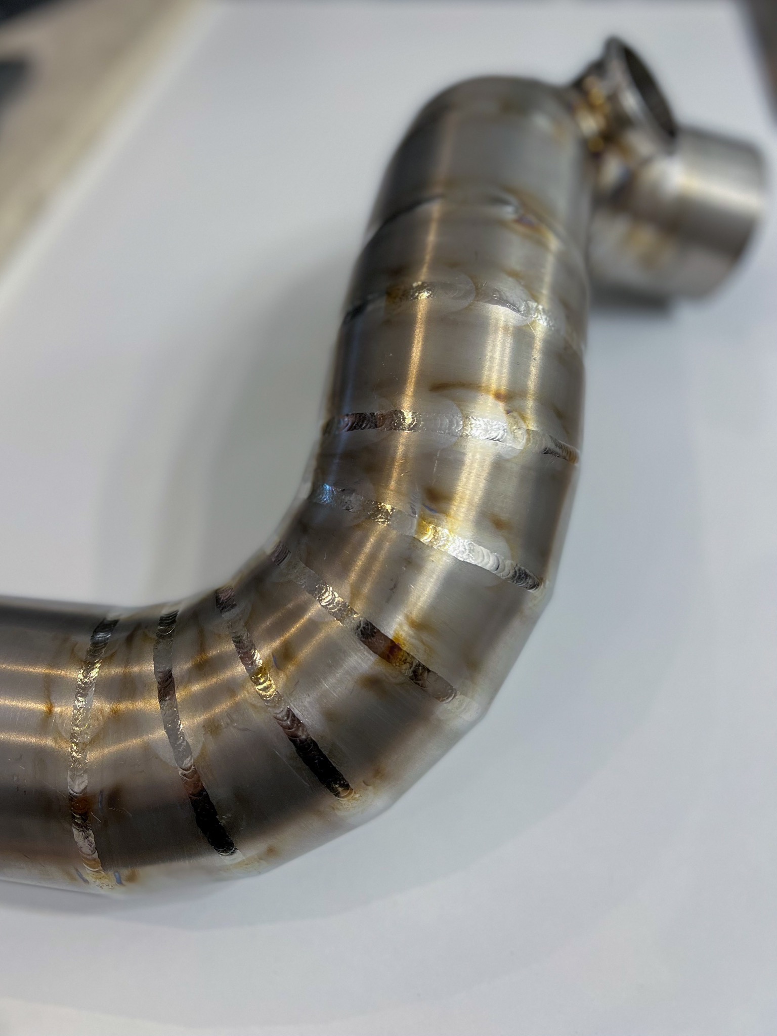 Precyzyjnie wykonane spawy na zakrzywionej rurze ze stali nierdzewnej, widoczne regularne ściegi spawalnicze z charakterystycznym wzorem, ułożone na białym tle.