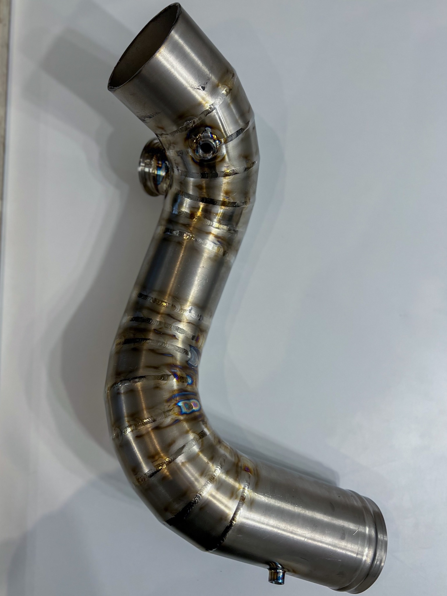 Zakrzywiona, spawana rura ze stali nierdzewnej z widocznymi śladami spawania i charakterystycznymi przebarwieniami termicznymi na spoinach, leżąca na białym tle.