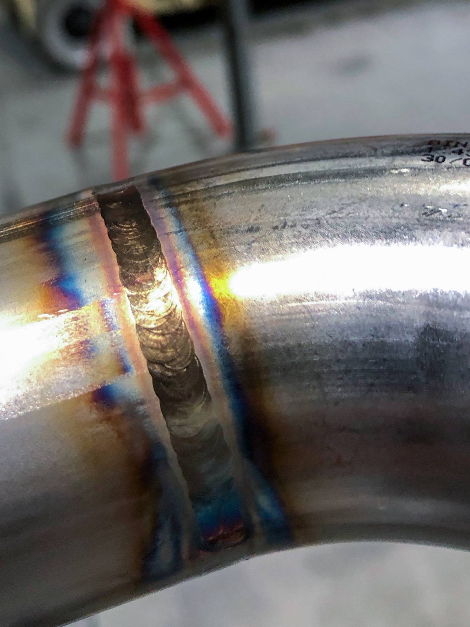 Szczegółowe ujęcie spoiny na metalowej rurze, z widocznymi tęczowymi przebarwieniami termicznymi i numerem seryjnym, w tle czerwony stojak warsztatowy.
