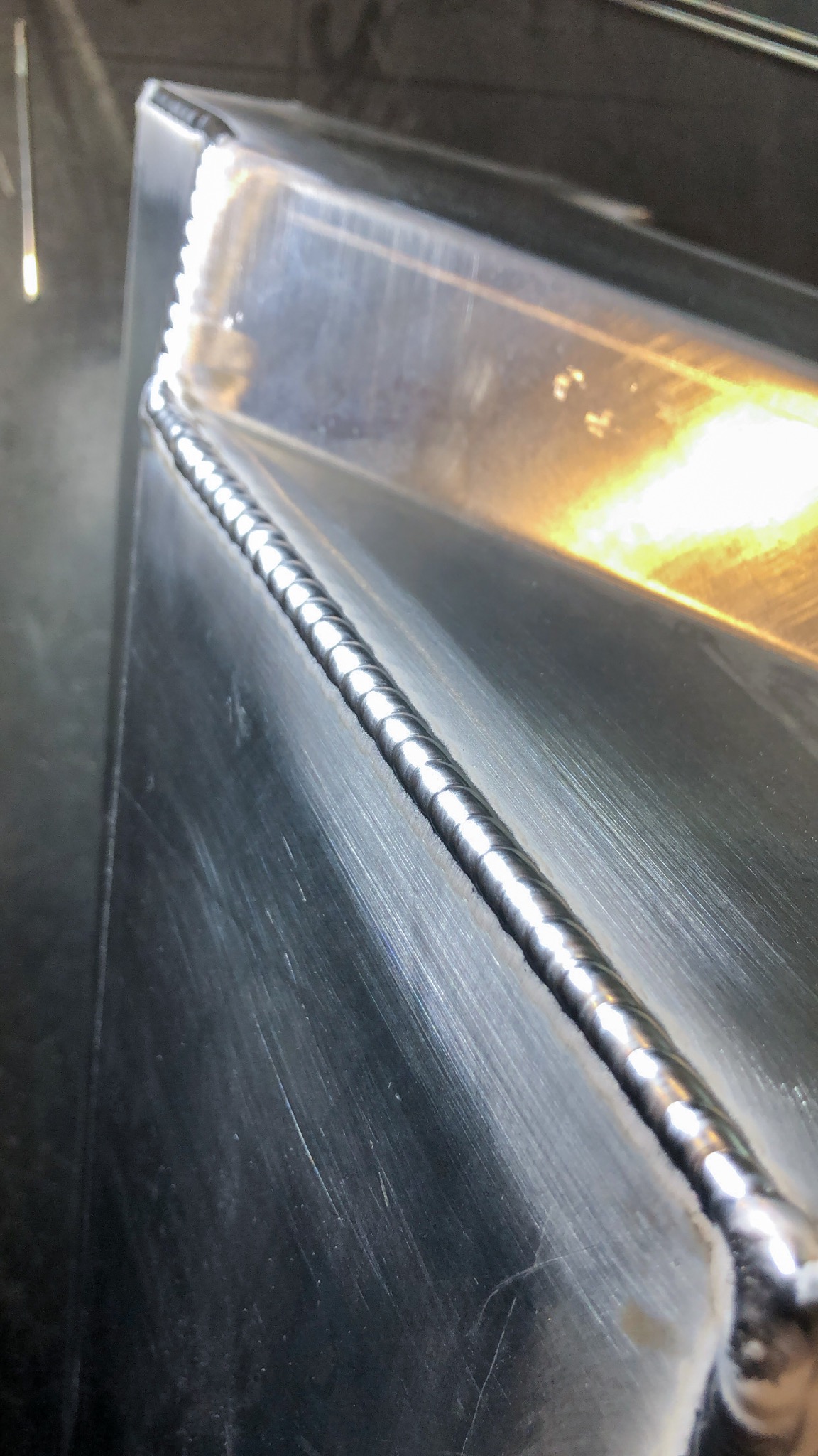 Szlifowana powierzchnia metalowa z precyzyjnym, równym spoiwem spawalniczym biegnącym wzdłuż krawędzi, widoczne delikatne rysy na metalu i odbicie światła.