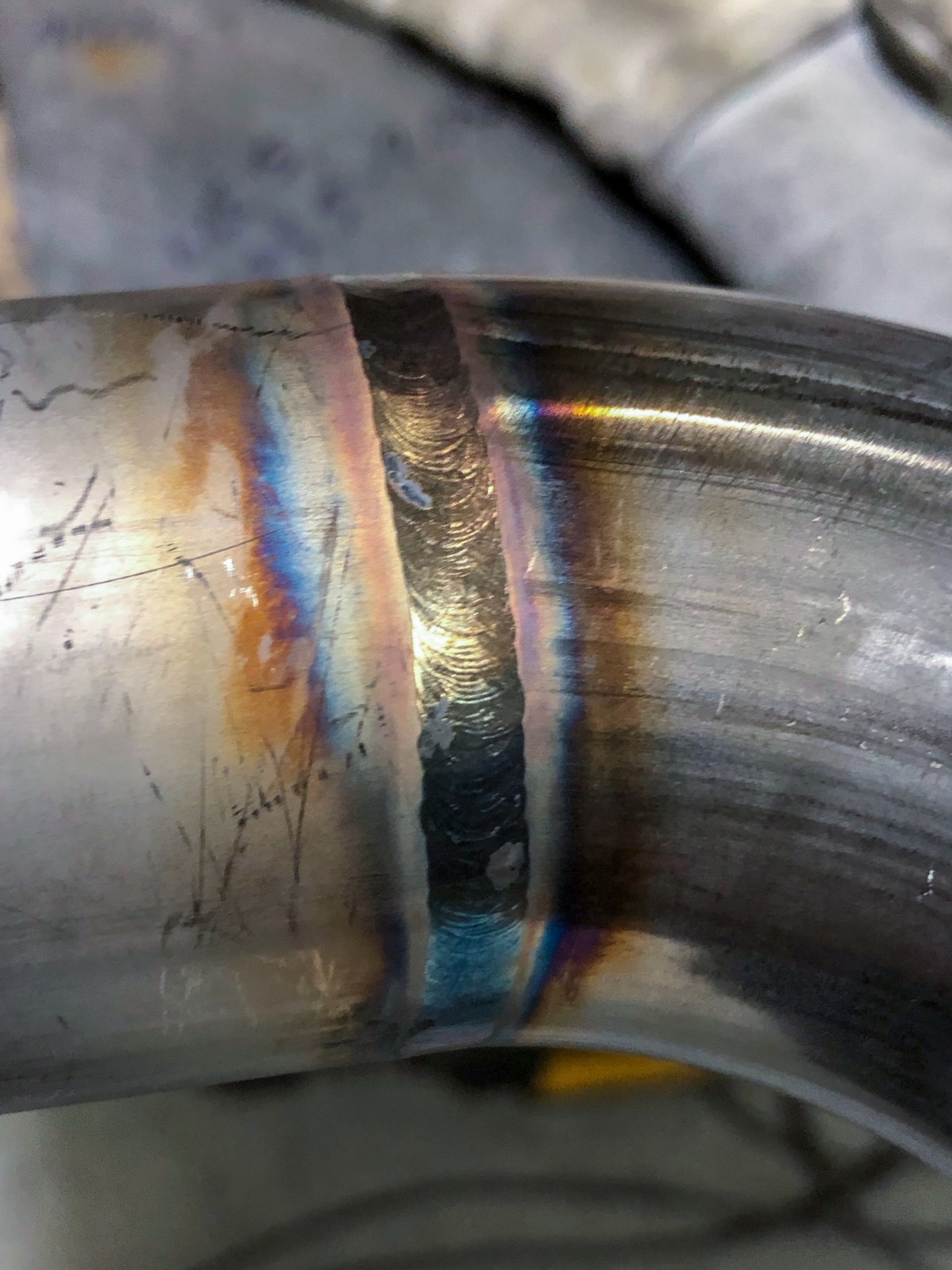 Szczegółowe zbliżenie spoiny na rurze ze stali nierdzewnej, widoczne tęczowe przebarwienia termiczne i delikatne ślady obróbki.