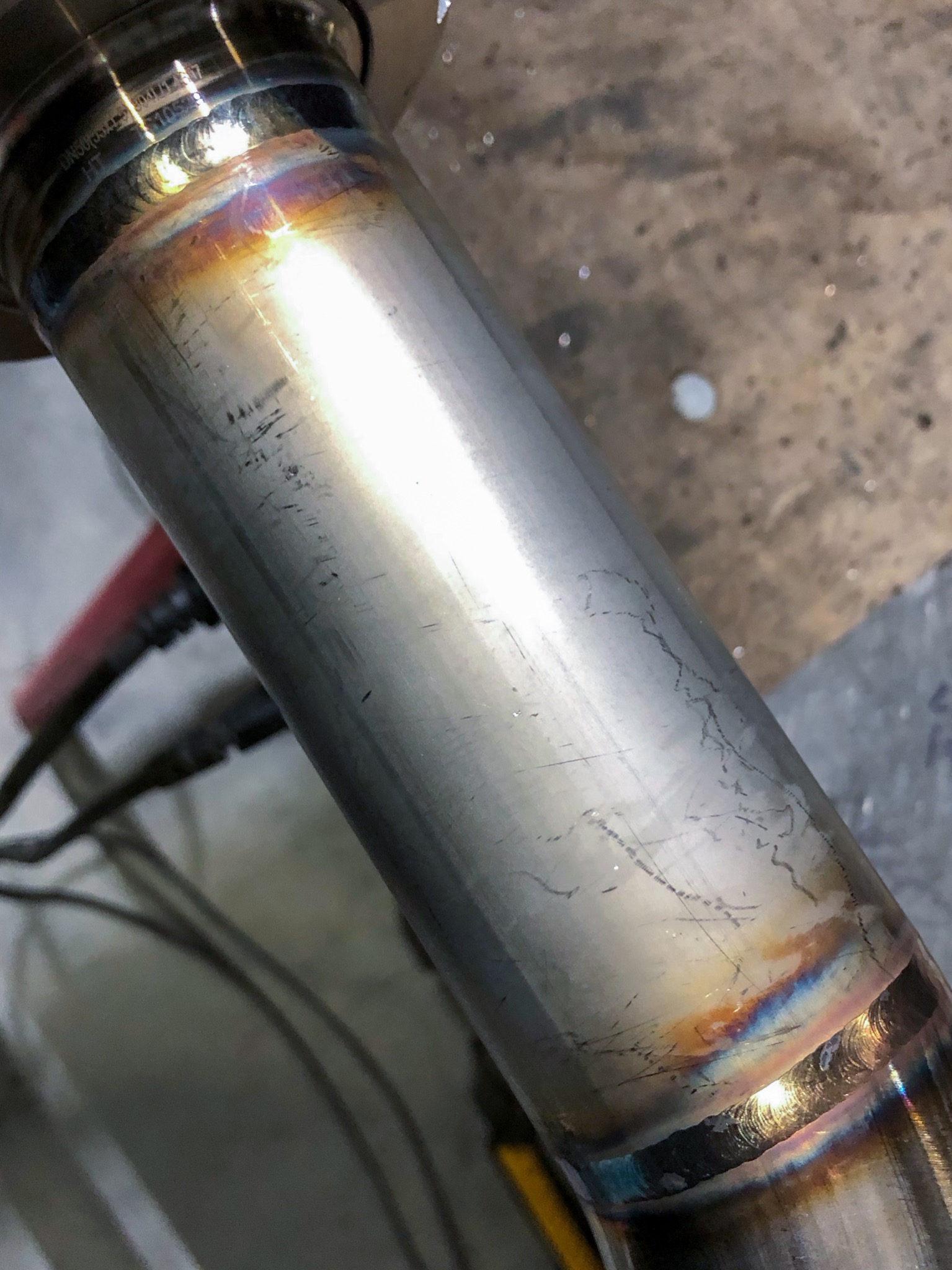 Błyszcząca, metalowa rura ze śladami spawania, widoczne przebarwienia termiczne na spoinie, drobne rysy i niedoskonałości na powierzchni.