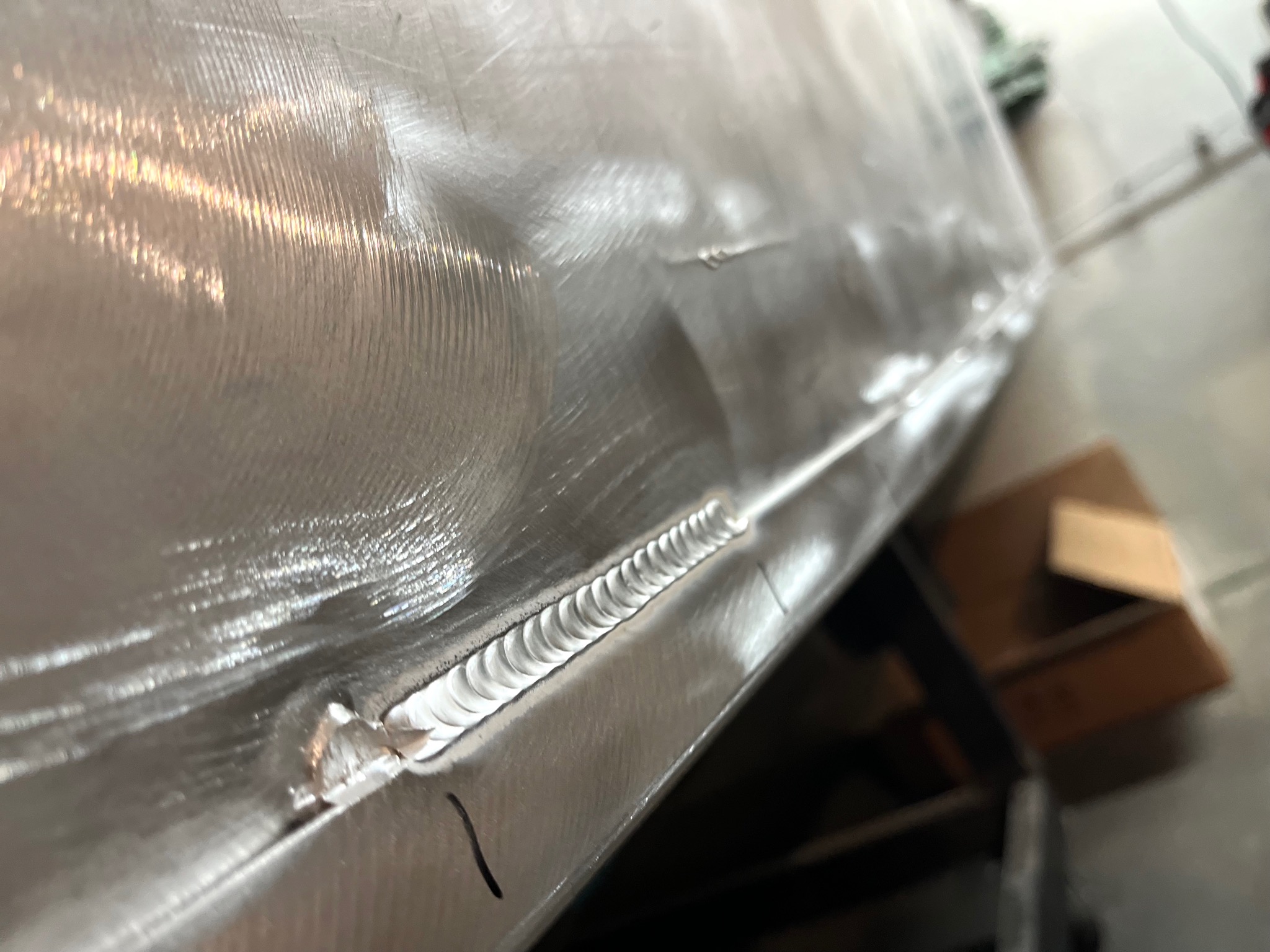 Szlifowana powierzchnia metalowa ze świeżo wykonanym spoiwem w postaci regularnych, drobnych łusek, widoczne ślady obróbki mechanicznej i drobne niedoskonałości.