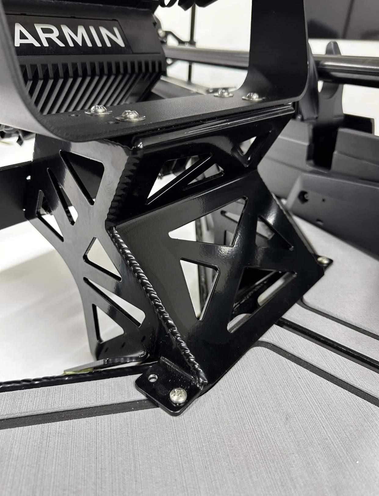 Czarny, metalowy wspornik z wyciętymi trójkątami, przymocowany śrubami do szarej powierzchni z czarnymi paskami, widoczne spawy i logo producenta 'ARMIN'.