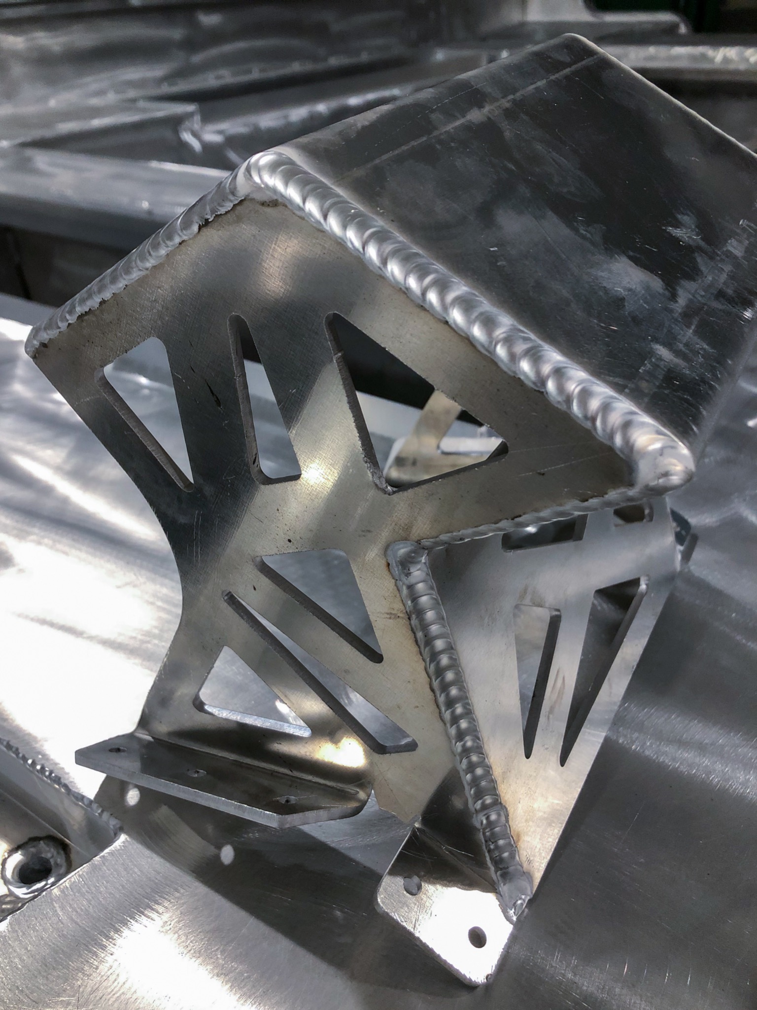 Metalowy element konstrukcyjny z widocznymi spawami i trójkątnymi wycięciami, leżący na metalowej powierzchni.