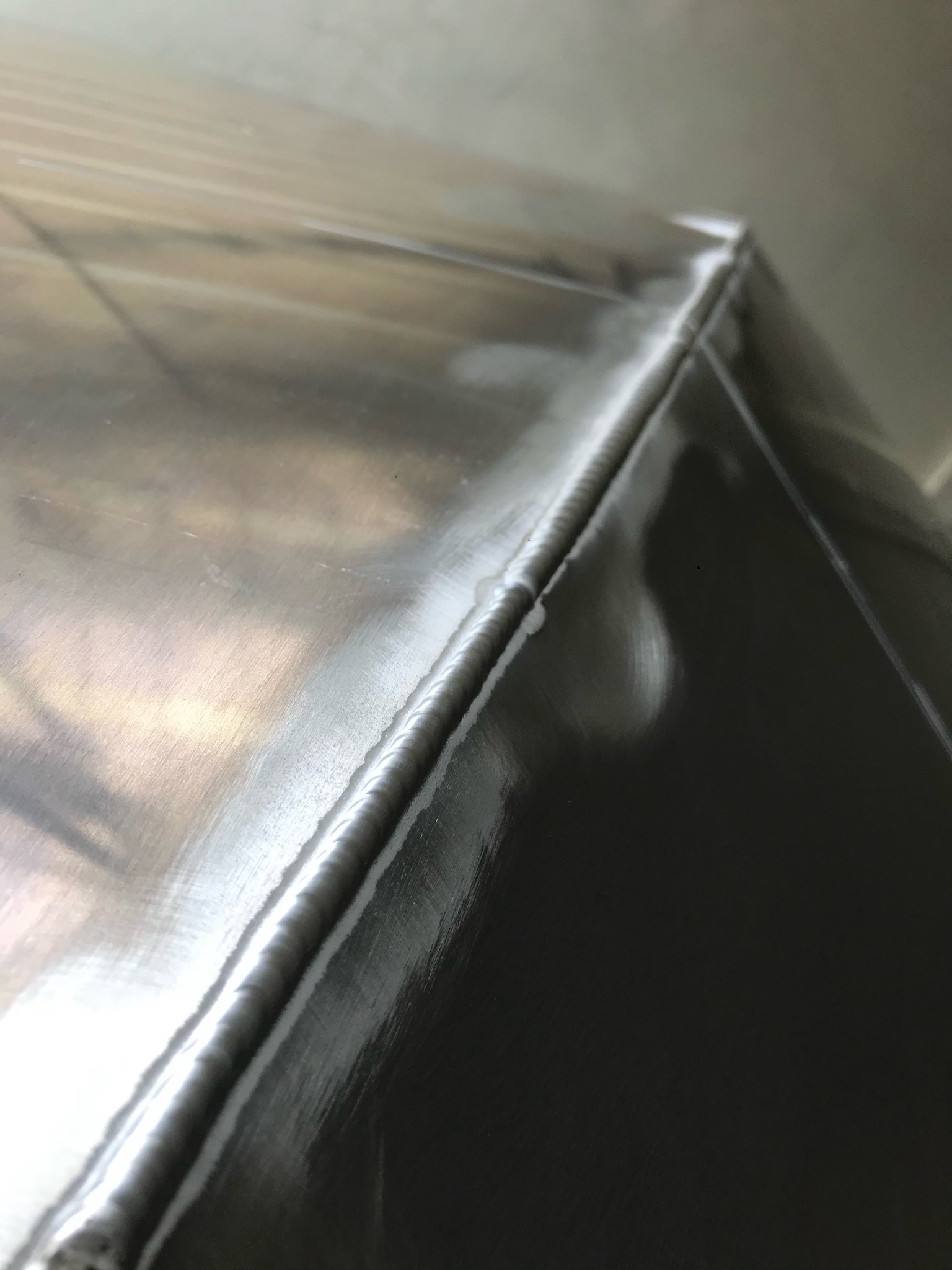 Szczegółowe zbliżenie na spawany narożnik metalowej konstrukcji; widoczny precyzyjny spaw, łączący dwie powierzchnie pod kątem prostym, z delikatnymi śladami szlifowania na powierzchni metalu.