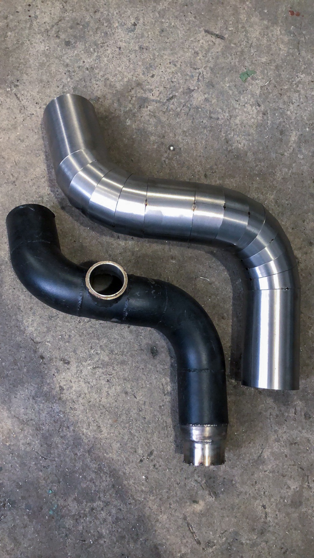 Dwie gięte rury: polerowana, segmentowa rura metalowa oraz czarna rura z przyspawanym kołnierzem, leżące na betonowej podłodze.