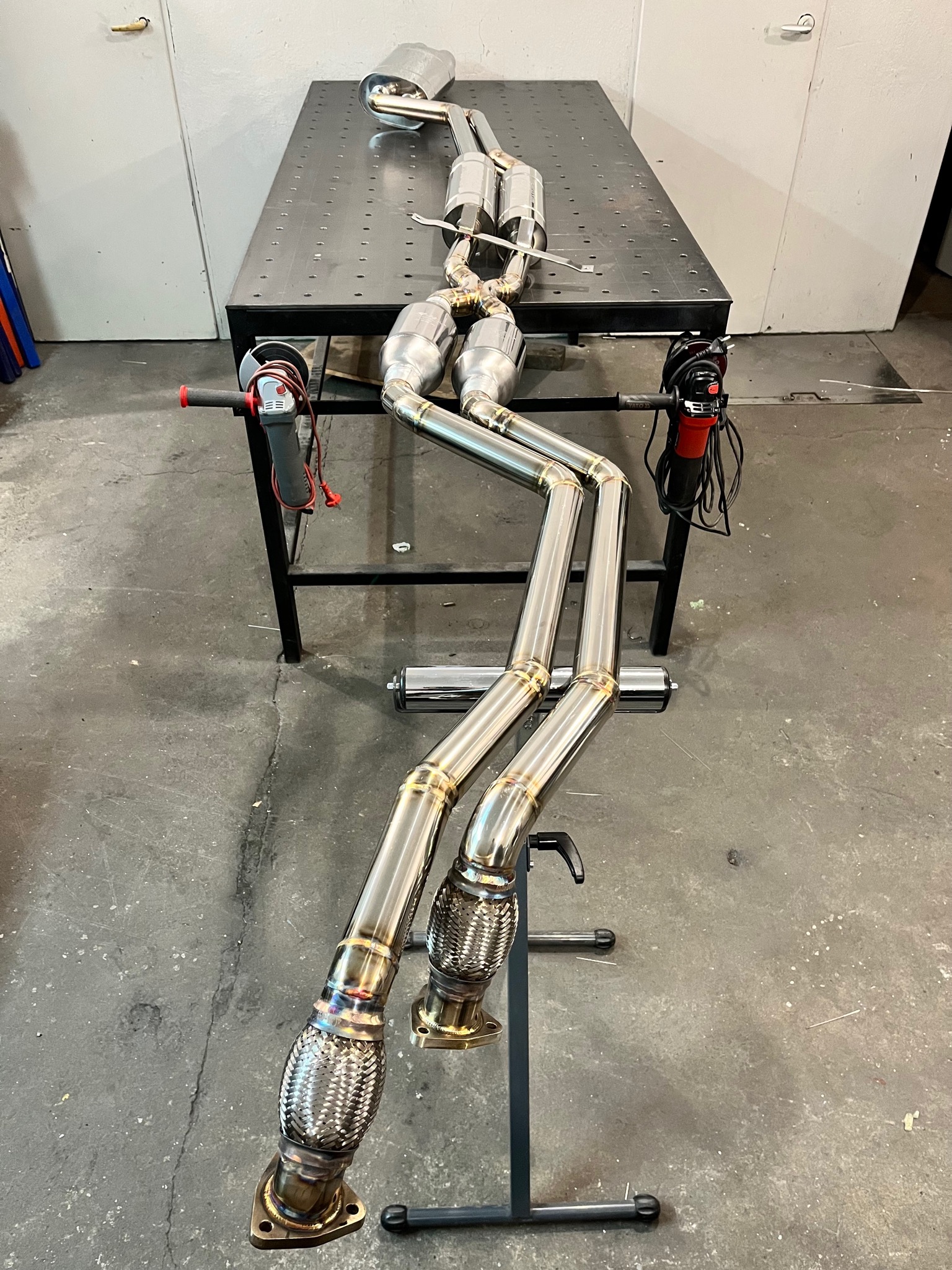 Polerowany, nierdzewny układ wydechowy z elastycznymi złączami i katalizatorami, leżący na perforowanym stole warsztatowym w pobliżu szlifierki kątowej.