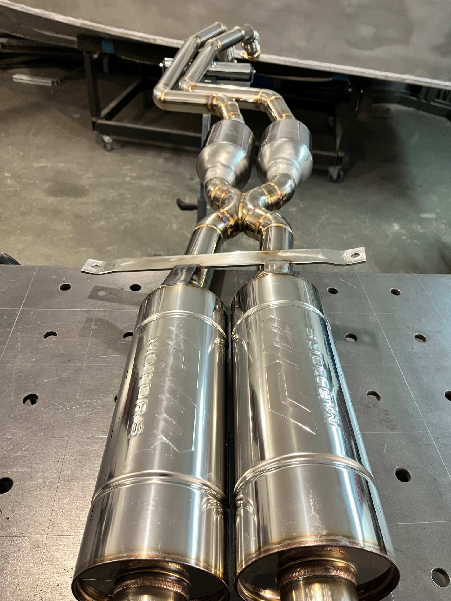 Kompletny układ wydechowy ze stali nierdzewnej z widocznymi spawami TIG, leżący na perforowanym stole spawalniczym.