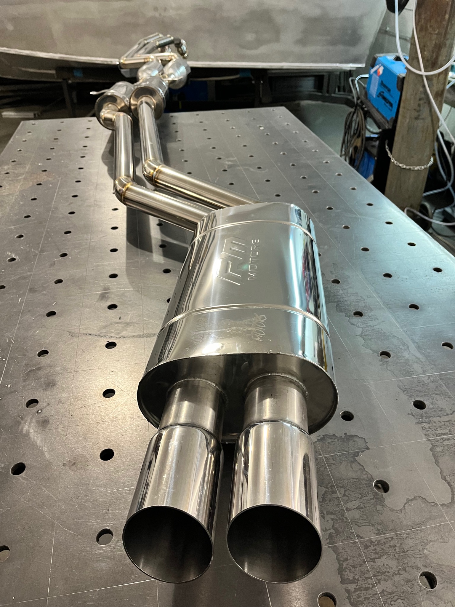 Polerowany, nierdzewny układ wydechowy z podwójną końcówką, leżący na perforowanym stole spawalniczym. Widoczne spawane łączenia i logo 'FM MOTORS' na tłumiku.