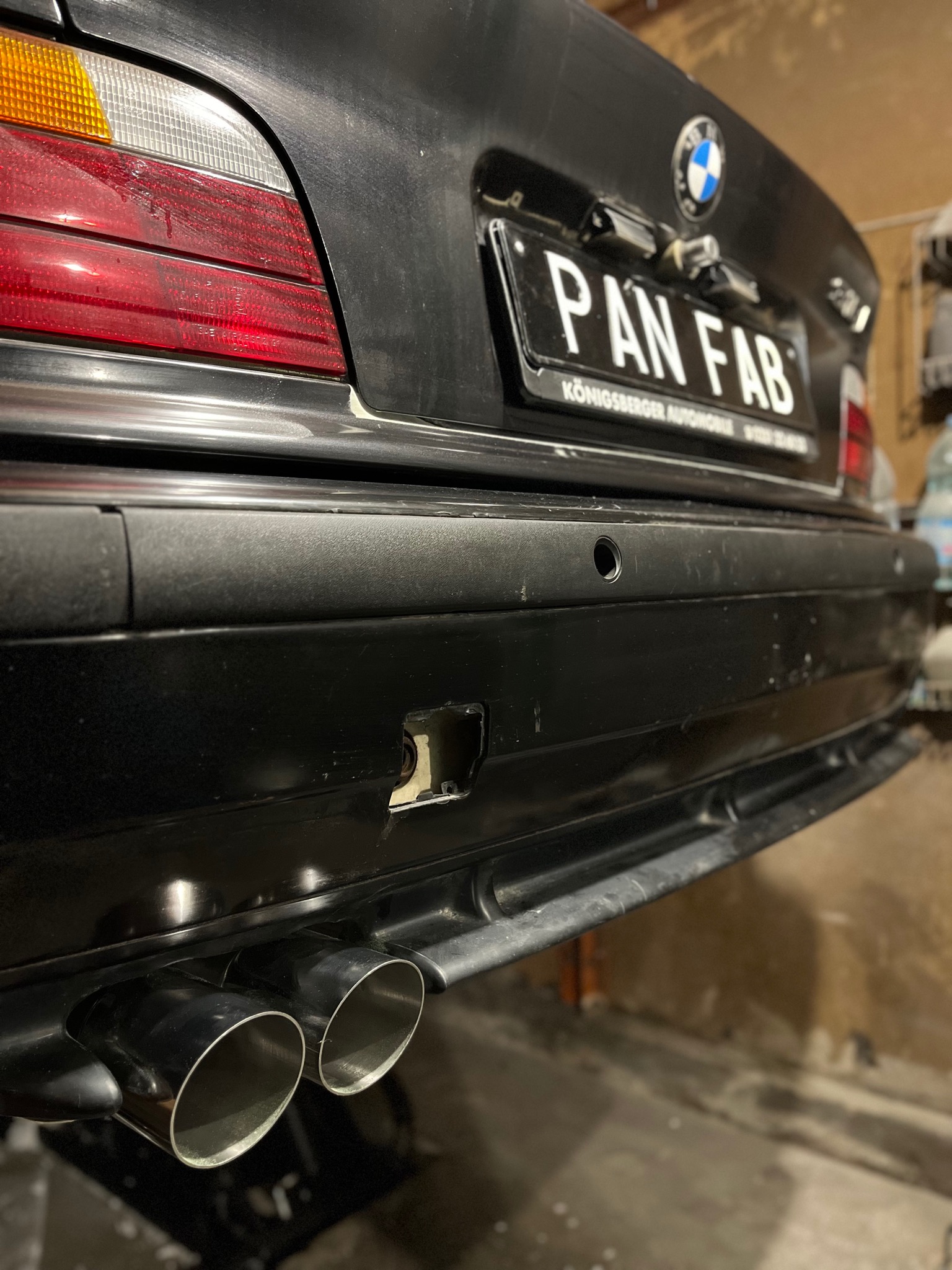 Czarny tylny zderzak BMW E36 z podwójnym chromowanym wydechem, widoczne ślady użytkowania na lakierze i brakujący element w zderzaku, tablica rejestracyjna z napisem 'PAN FAB'.