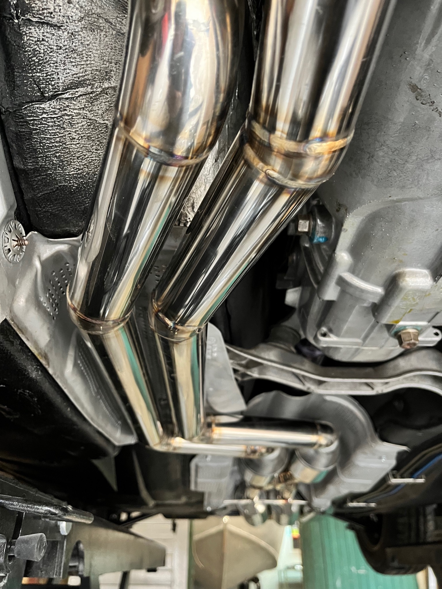 Widok od spodu na spawany, polerowany układ wydechowy z rur nierdzewnych, zamontowany w samochodzie, z widocznymi spawami i elementami mocującymi.