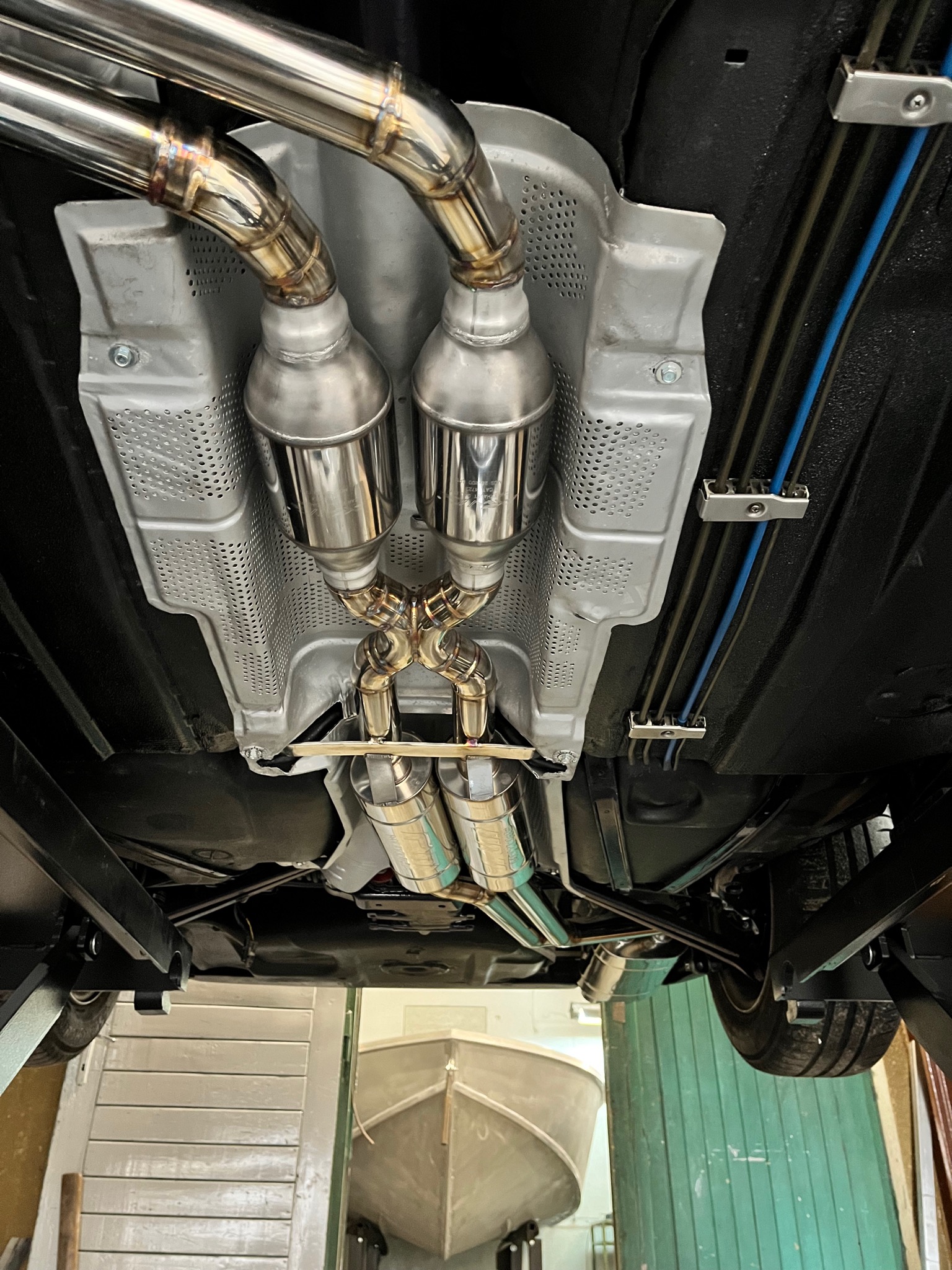 Widok od dołu na podwozie samochodu z zamontowanym, nowym układem wydechowym ze stali nierdzewnej. Widoczne spawane łączenia rur wydechowych oraz osłona termiczna z perforowanej blachy.