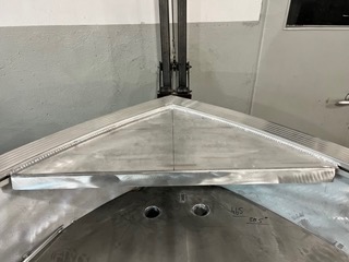 Metalowa konstrukcja łodzi w trakcie spawania, widoczne ślady po spawaniu, surowe wykończenie, pionowe wsporniki nad konstrukcją.