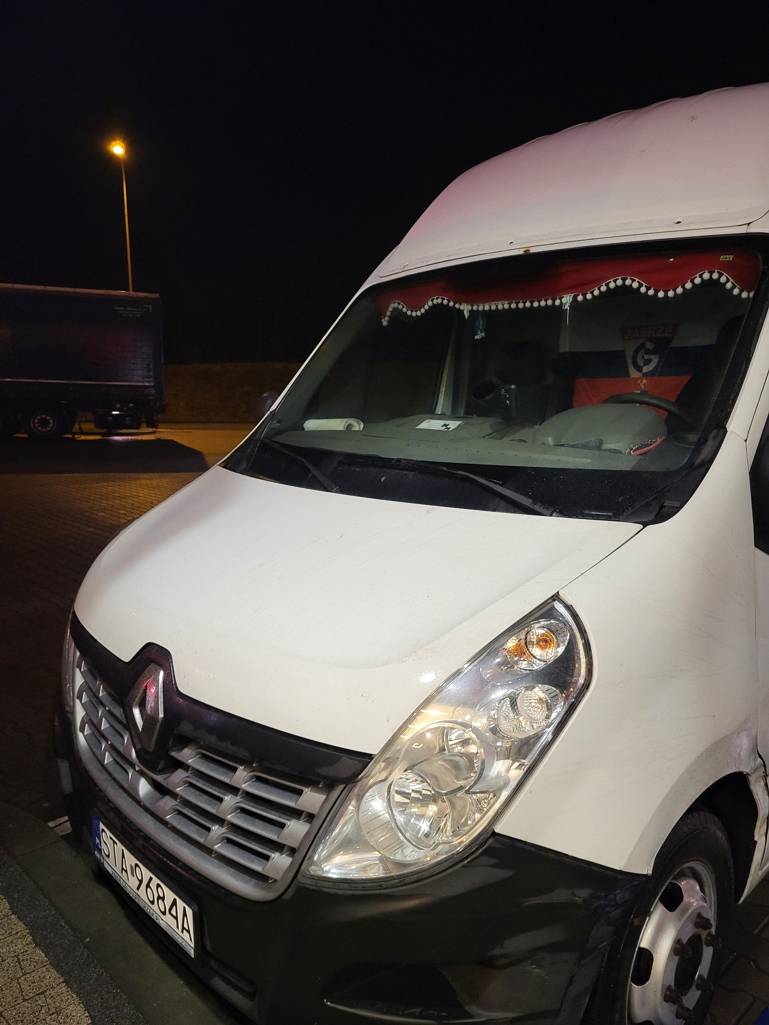 Biały bus Renault Master z flagą w oknie i czerwoną firanką z pomponami, zaparkowany nocą przy oświetlonej ulicy, widoczny fragment naczepy w tle.