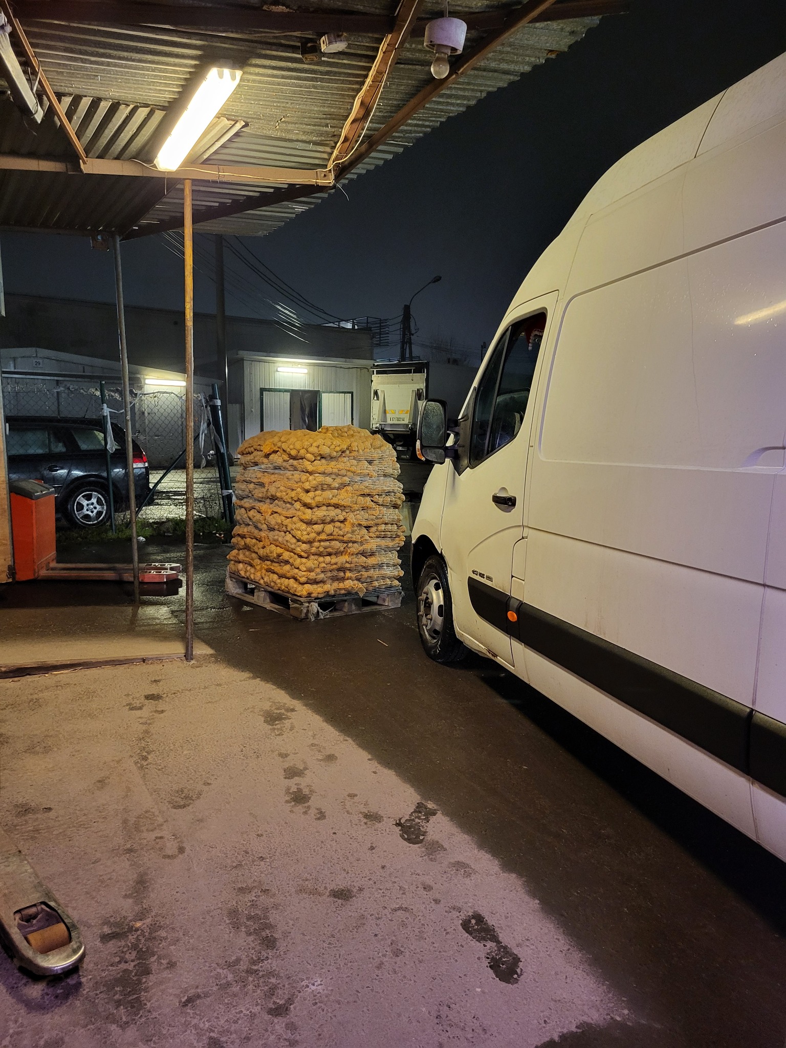 Biały bus dostawczy zaparkowany obok palety z workami z ziemniakami pod zadaszeniem, widoczne ślady na mokrej nawierzchni i oświetlenie lampą jarzeniową.