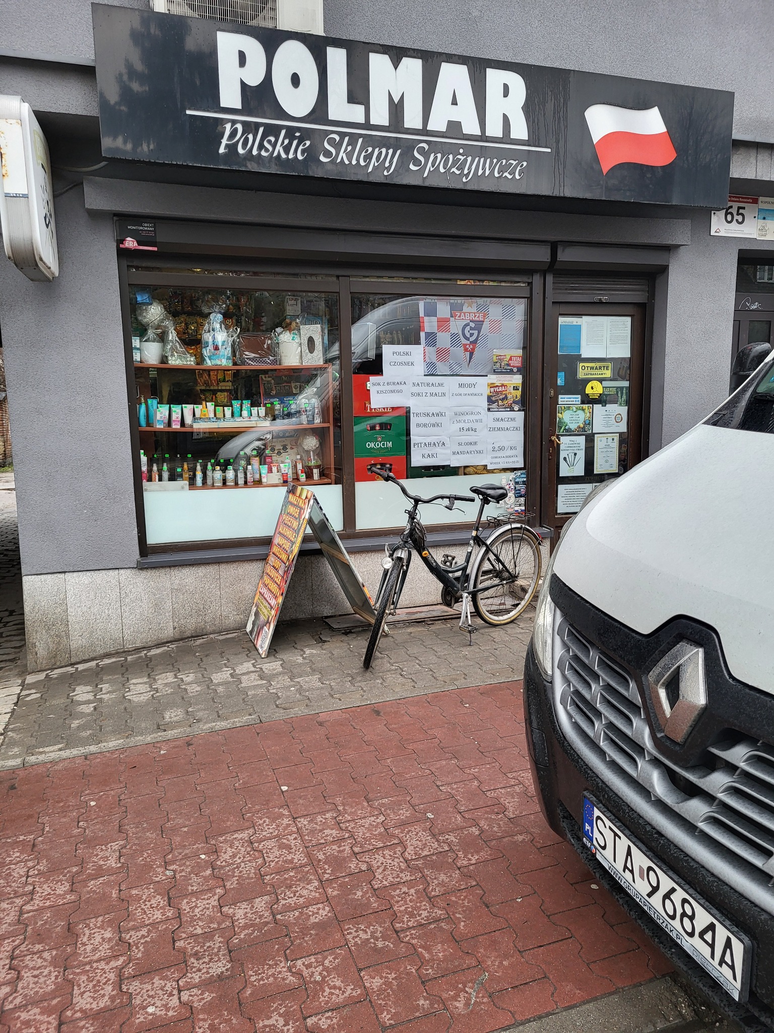 Fasada sklepu spożywczego 'POLMAR' z polskimi produktami, rower zaparkowany przed wejściem, fragment zaparkowanego białego busa widoczny z przodu