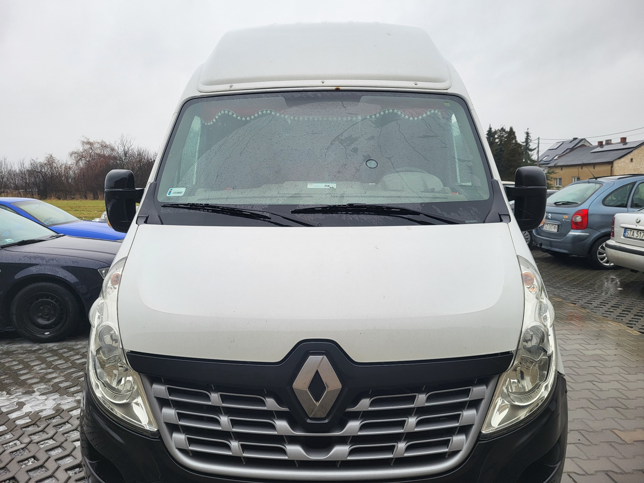 Przód białego busa Renault Master z dekoracyjną zasłonką na przedniej szybie, zaparkowanego na kostce brukowej w pochmurny dzień.