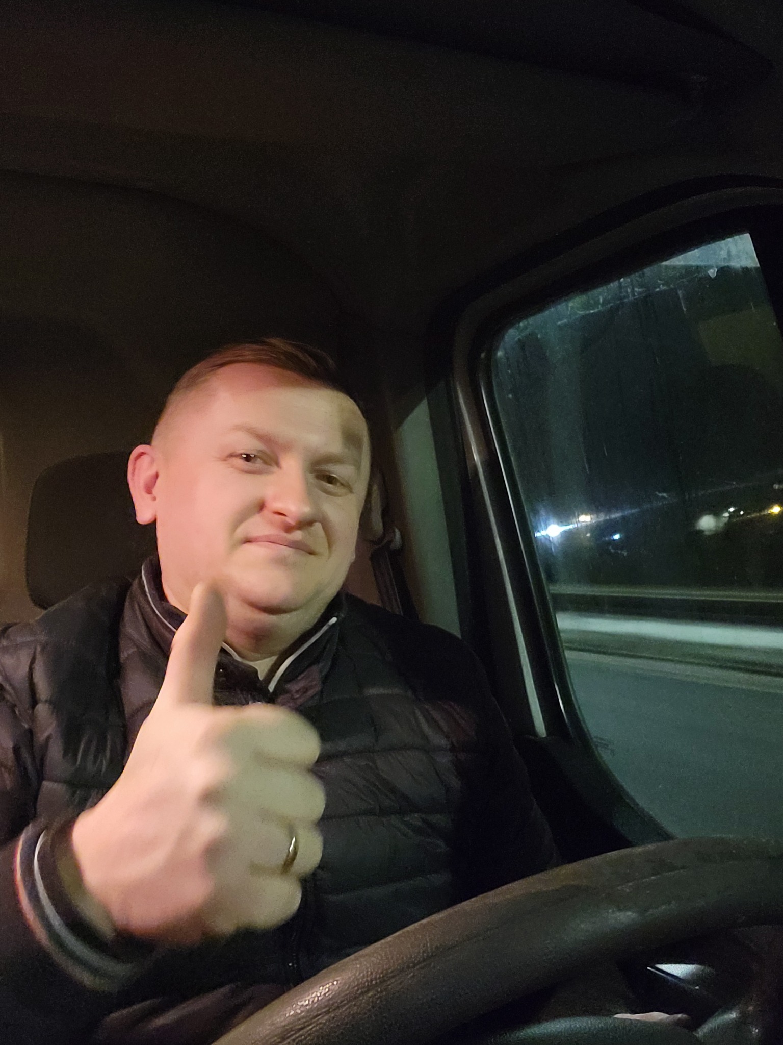 Uśmiechnięty kierowca w czarnej kurtce, z kciukiem w górę, siedzący za kierownicą busa w trakcie nocnej podróży.