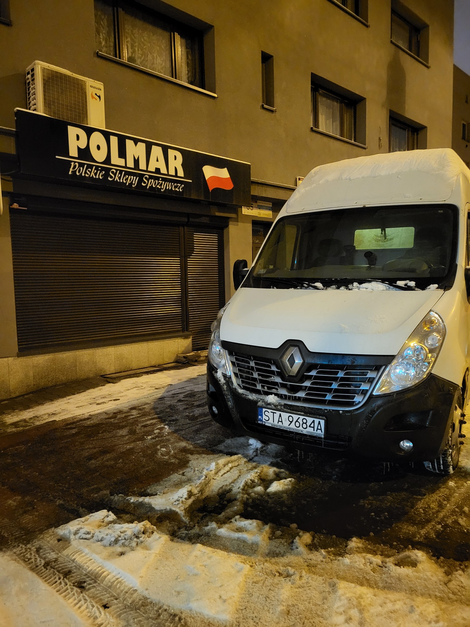 Biały bus Renault zaparkowany przed sklepem spożywczym 'POLMAR' w zimowy wieczór, z widocznymi śladami opon na śniegu.