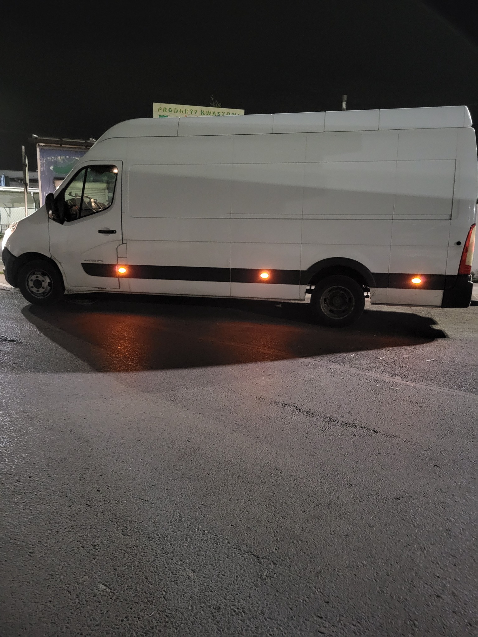 Biały bus dostawczy zaparkowany na asfalcie nocą, z włączonymi światłami pozycyjnymi, widoczny z boku na tle ciemnego nieba i tablicy reklamowej.