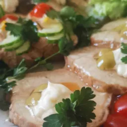 Dekoracyjnie podane plastry pieczeni z sosem, pomidorkami cherry, og&oacute;rkiem i zieloną sałatą na białym talerzu, z bliska.