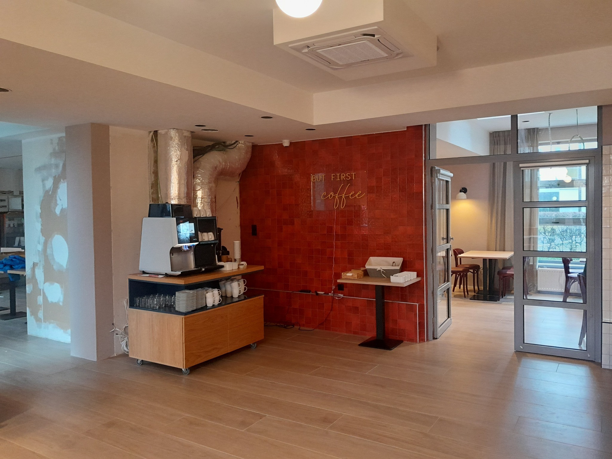 Wnętrze biura z aneksem kuchennym, widoczna ekspres do kawy, czerwona ściana z napisem neonowym 'BUT FIRST coffee', przeszklone drzwi do sali konferencyjnej oraz fragment ściany w trakcie...