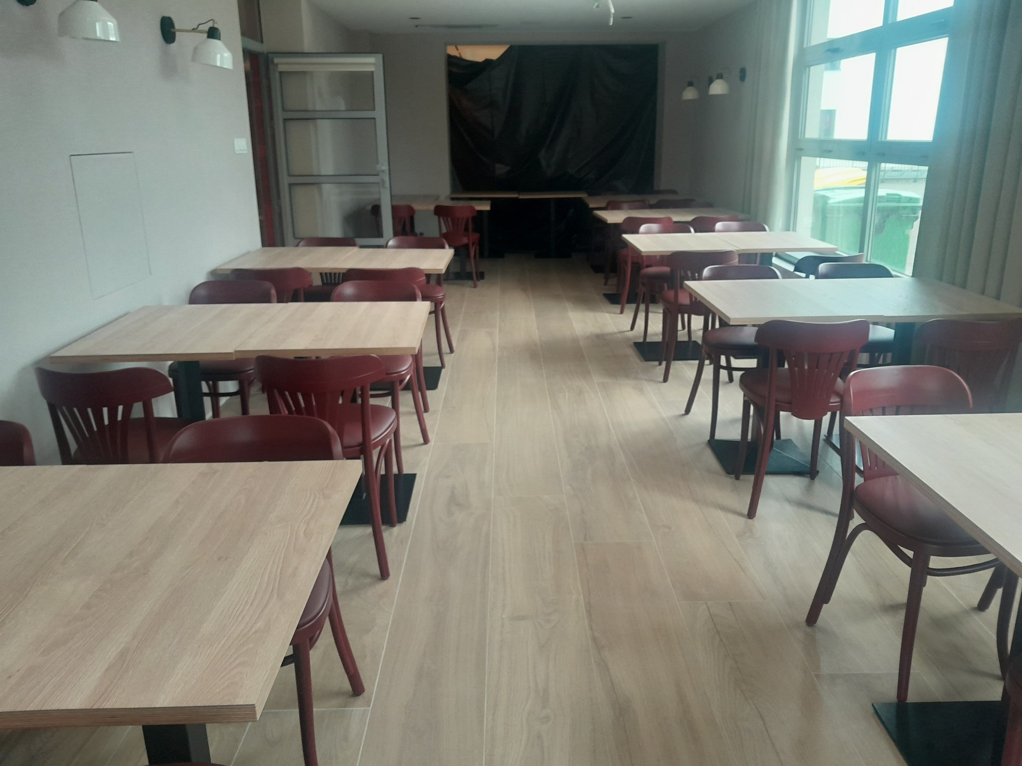 Sala restauracyjna z drewnianymi stołami, krzesłami w kolorze czerwonego wina i podłogą z paneli, zasłonięte okno w tle.