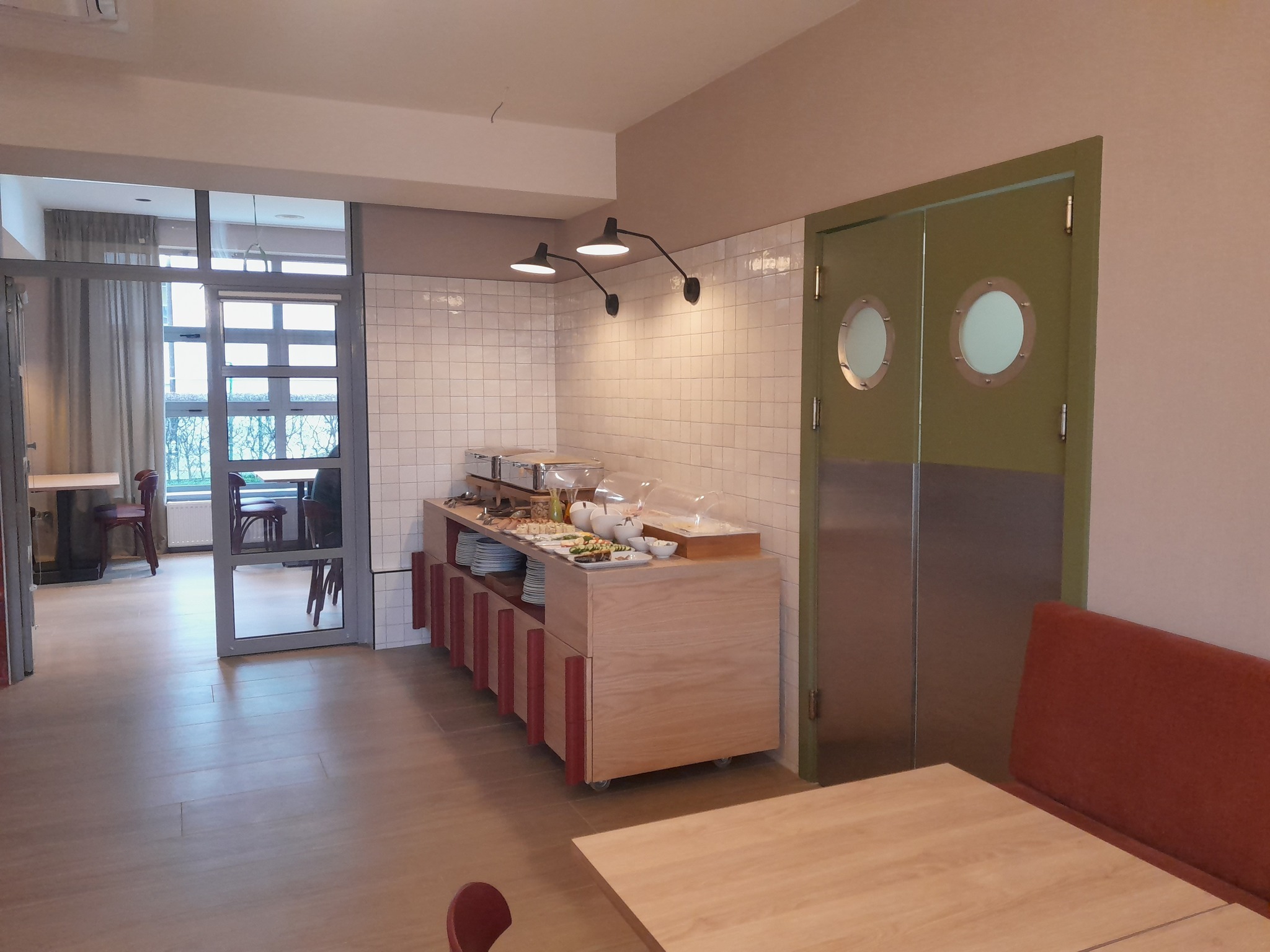 Sala restauracyjna z bufetem śniadaniowym: drewniany stół z czerwoną kanapą, bufet z potrawami, płytki na ścianach i drzwi z okrągłymi okienkami.