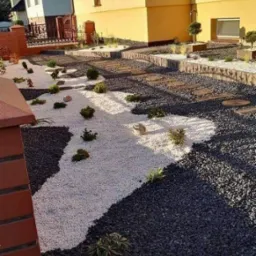 Ogród z dekoracyjnymi białymi i czarnymi kamieniami, drewnianymi stopniami i niskimi roślinami ozdobnymi, widok z góry na tle domu.