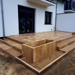 Topmates - Nowoczesny taras z kostki brukowej w odcieniach brązu, schody z kostki prowadzące do domu, donice z kostki wypełnione piaskiem, widok z poziomu gruntu.