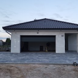 Topmates - Nowo ułożona, szara kostka brukowa przed nowoczesnym domem z otwartym garażem, wewnątrz widoczny pracownik z zagęszczarką. Obrzeża chodnikowe oddzielają kostkę od ziemi. Częściowo widoczna...