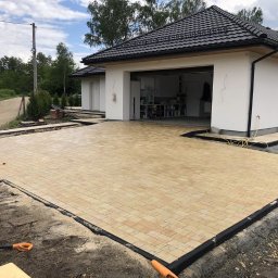 Topmates - Nowo ułożony, beżowy podjazd z kostki brukowej przed nowoczesnym domem z białymi ścianami i czarnym dachem, widoczne narzędzia i obrzeża, prace w toku.