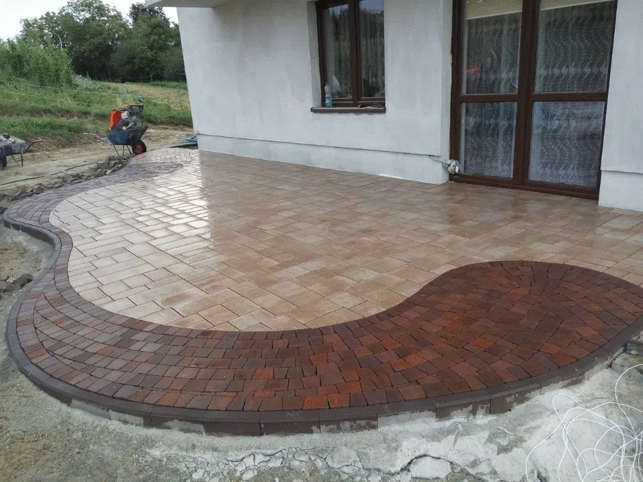 Ułożona kostka brukowa w kształcie fali z obrzeżem z ciemniejszej kostki przy domu, widoczne narzędzia i niedokończone prace ziemne.