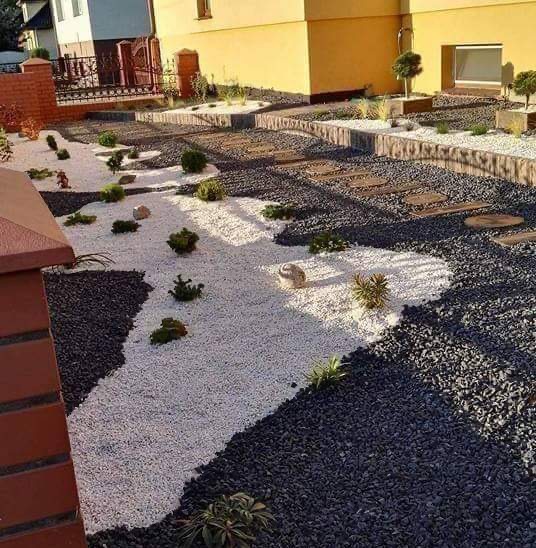 Ogród z dekoracyjnymi białymi i czarnymi kamieniami, drewnianymi stopniami i niskimi roślinami ozdobnymi, widok z góry na tle domu.