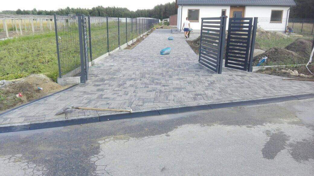 Ułożona szara kostka brukowa prowadząca do domu z otwartą, nowoczesną bramą; widoczne narzędzie brukarskie i worki z piaskiem.
