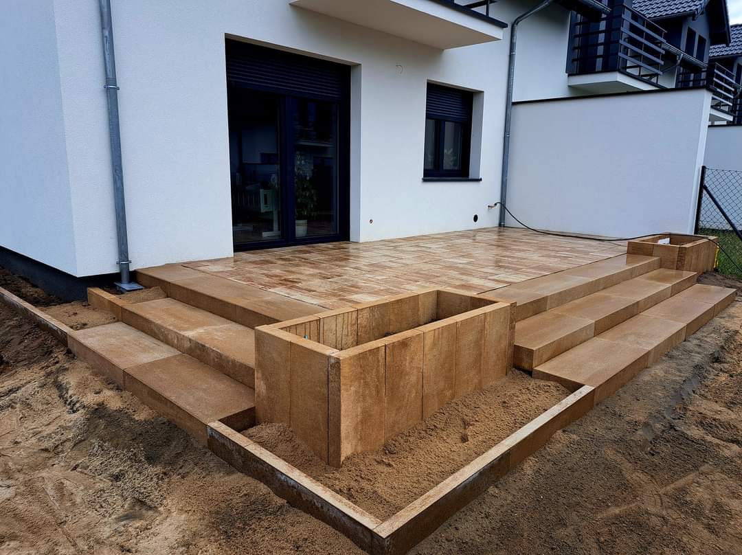 Nowoczesny taras z kostki brukowej w odcieniach brązu, schody z kostki prowadzące do domu, donice z kostki wypełnione piaskiem, widok z poziomu gruntu.