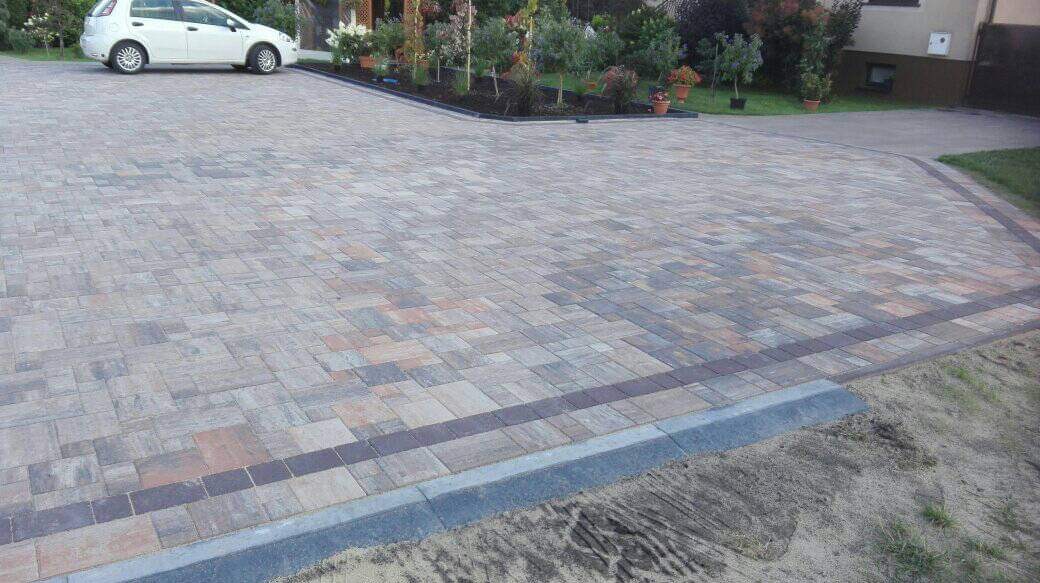 Ułożona nawierzchnia z kostki brukowej o nieregularnym wzorze i zróżnicowanej kolorystyce, obrzeża z ciemniejszej kostki, widoczny fragment trawnika i zaparkowany biały samochód.