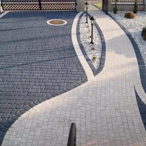 Ułożona kostka brukowa w dwóch kolorach, szary i jasnoszary, tworząca ścieżkę i podjazd z ozdobnym pasem białych kamieni i niskimi lampami wzdłuż ścieżki, widoczny właz kanalizacyjny.