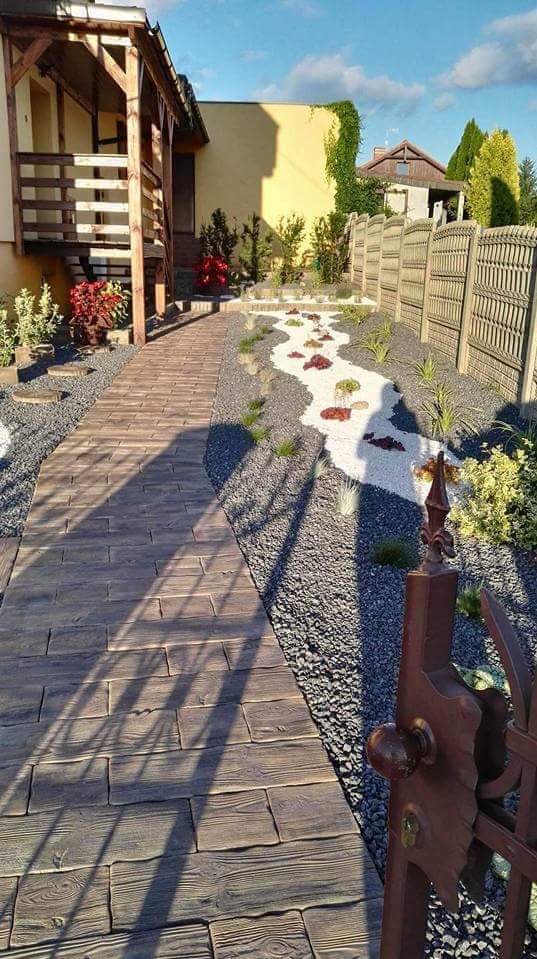 Ogród z drewnopodobną ścieżką z kostki, szarym żwirem i dekoracyjnym korytem z białych kamieni i czerwonych akcentów, widok zza ozdobnej bramy.