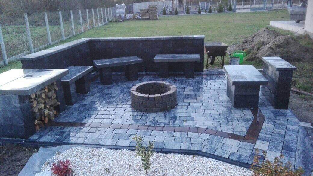 Zewnętrzna strefa relaksu z paleniskiem otoczona kostką brukową w odcieniach szarości i grafitu, z ławkami i miejscem na drewno, widok na trawnik i ogrodzenie.