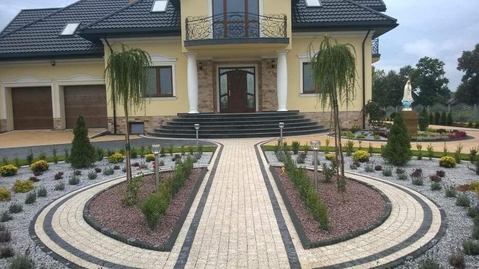 Elegancki podjazd i chodnik z kostki brukowej w odcieniach beżu i szarości, prowadzący do okazałego domu z kolumnami, otoczony rabatami z krzewami i ozdobnym żwirem, z widoczną figurą Matki Boskiej...