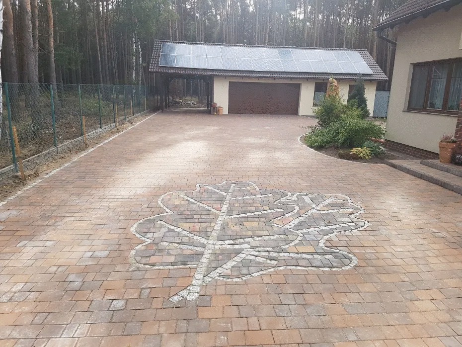 Szeroki podjazd z kostki brukowej w odcieniach czerwieni i brązu, z dekoracyjnym motywem liścia dębu wykonanym z jasnej kostki. W tle dom z panelami słonecznymi i las.
