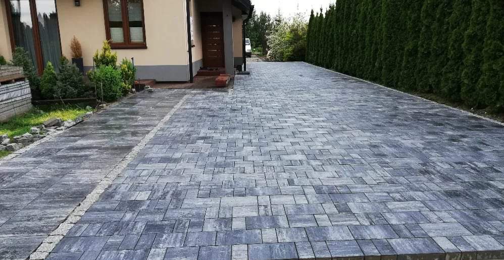 Nowoczesna, szara kostka brukowa ułożona na podjeździe w Gnieźnie, z widoczną linią oddzielającą od chodnika i zielonym żywopłotem w tle.