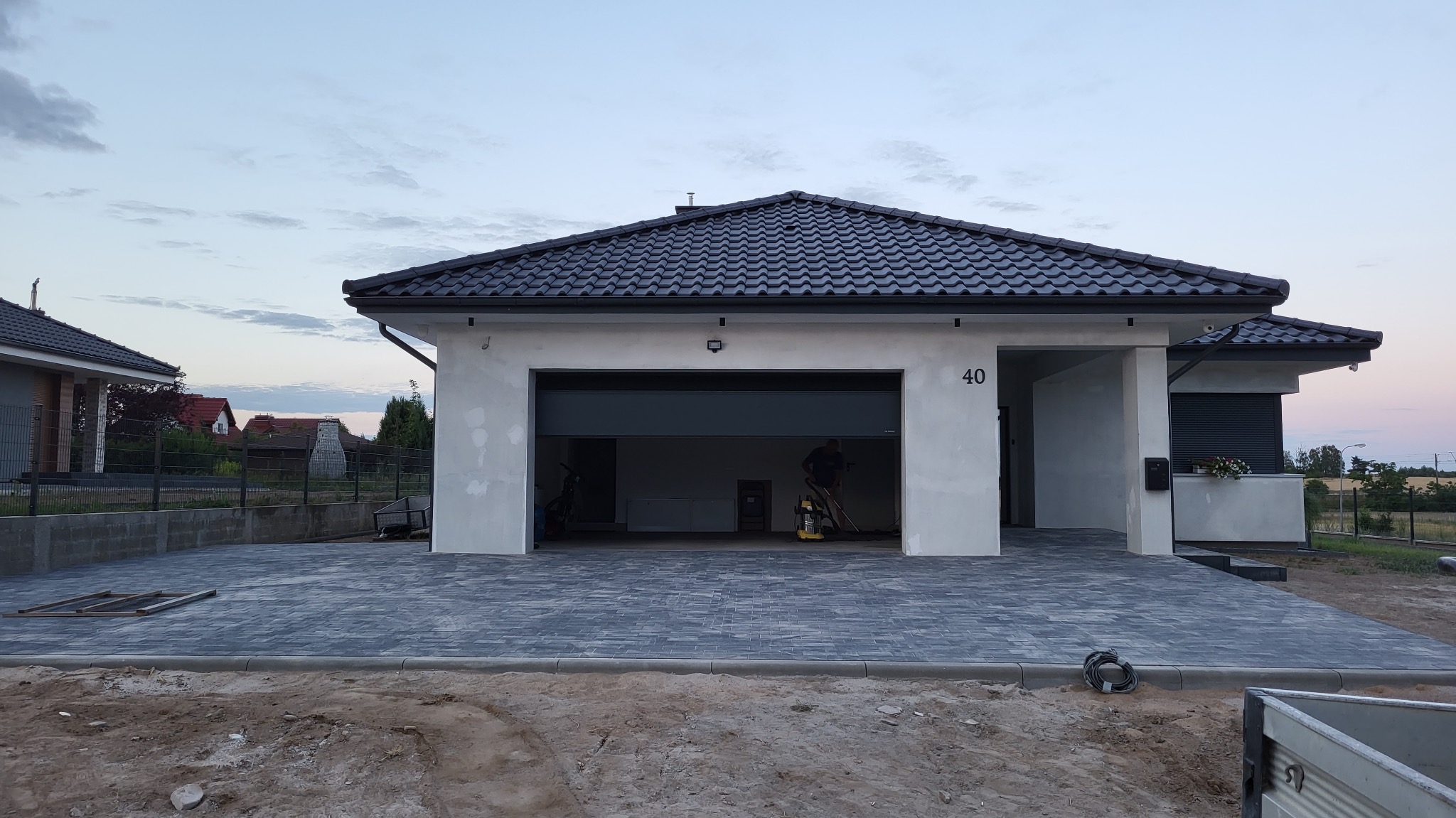 Nowo ułożona, szara kostka brukowa przed nowoczesnym domem z otwartym garażem, wewnątrz widoczny pracownik z zagęszczarką. Obrzeża chodnikowe oddzielają kostkę od ziemi. Częściowo widoczna...